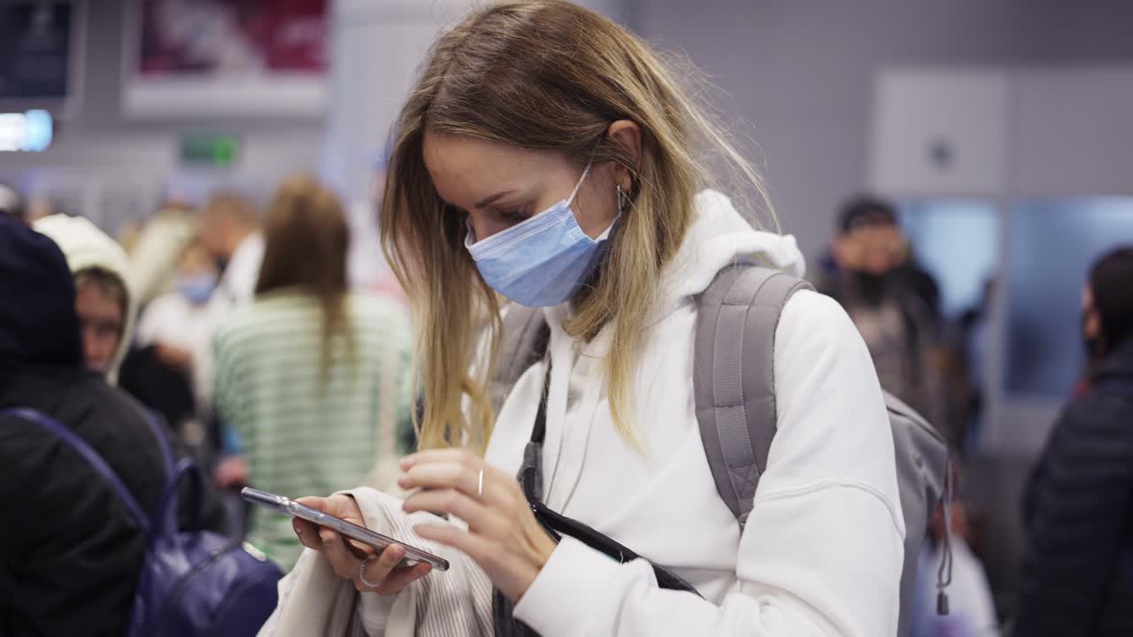 mujer con máscara usando un teléfono inteligente mientras espera un vuelo en la multitud del aeropuerto