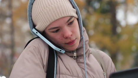 close view of light skin lady dressed in beige jacket and knitted cap talking on mobile phone with blue case outdoors during cold season, blurred background of autumn trees and natural soft light