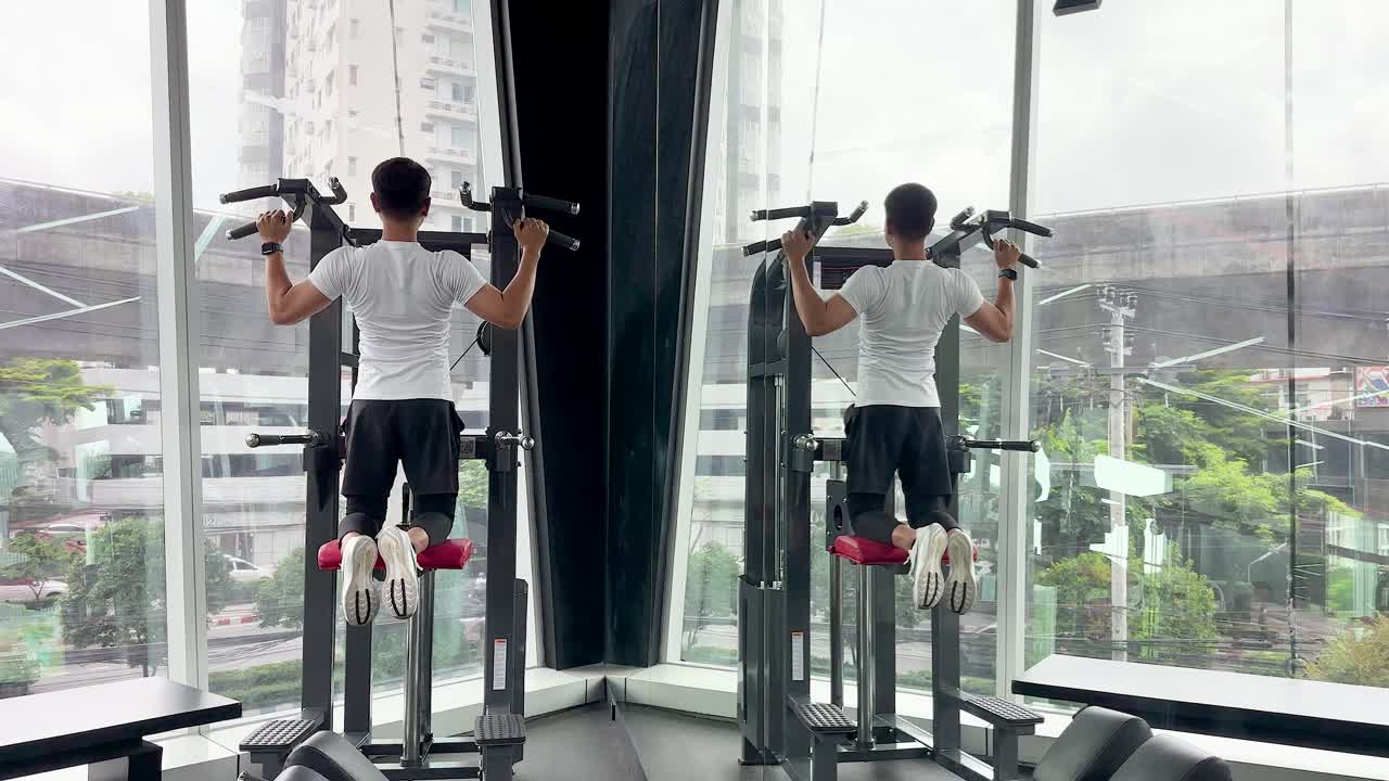 Athletic man uses assisted pull-up machine in bright gym, large windows reveal urban cityscape outside