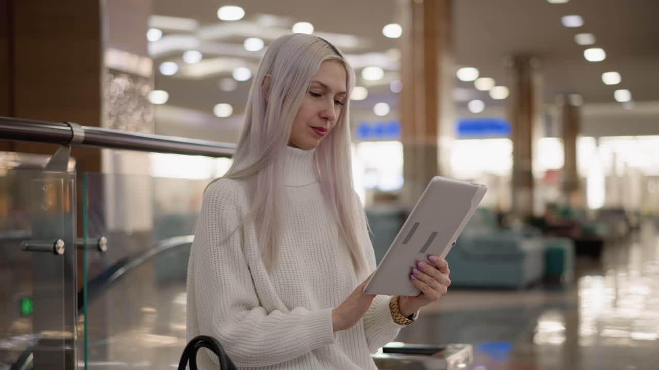 expectant mother seated on bench in bright mall hall, wearing white sweater and jeans, engrossed in tapping tablet, black handbag at side, modern interior lighting and glass railing backdrop