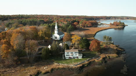 Aerial-shot-of-white-church-and-houses-by-a-river-in-autumn-landscape,-Phippsburg-Maine