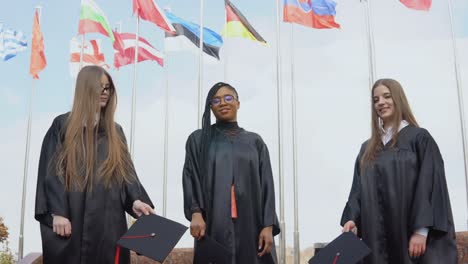 three young women of different nationalities joyfully throw hats up against many flags of different countries fluttering in the wind on the background. outdoor view