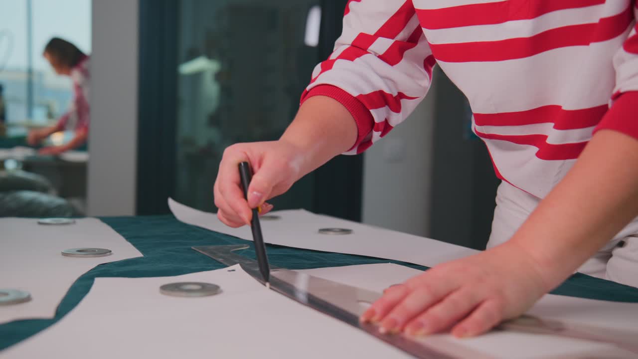 Person marks fabric with precision tool. Hands steady, focused on cutting pattern. Background shows modern office space. Ideal for fashion design, tailoring, or crafting projects