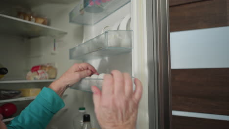 elderly woman in green sweatshirt reaching into refrigerator to grab eggs from top shelf. modern kitchen setting with organized food items and soft lighting, focusing on daily cooking routine