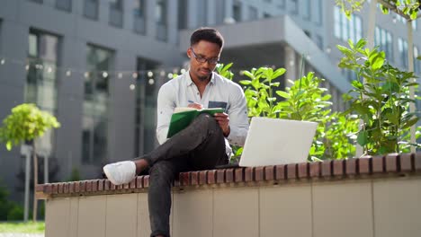 young african american male student sitting in a city park on a bench with laptop and notebook studying online outdoors