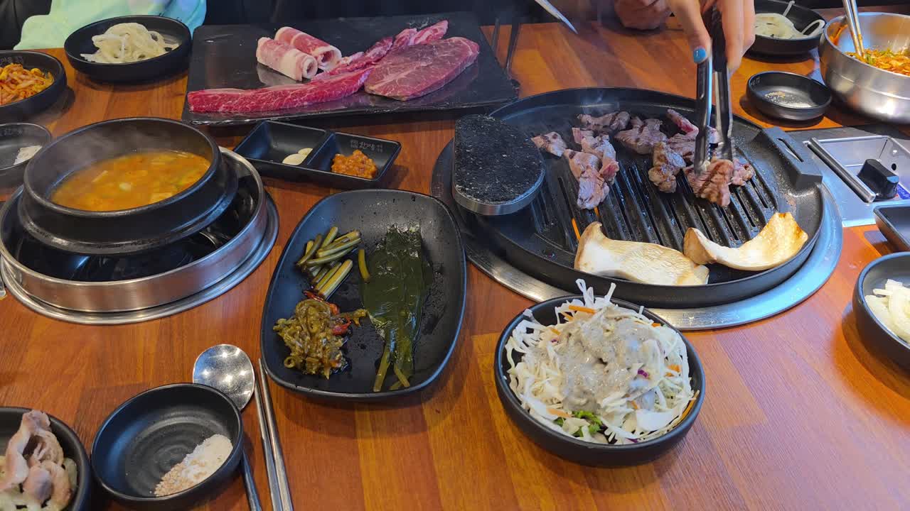 Hands grilling and cutting marbled beef and king oyster mushrooms on a tabletop grill with assorted Korean side dishes - top view