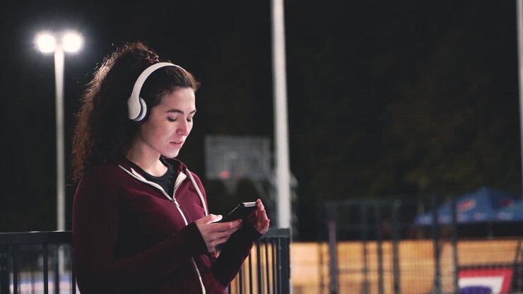 Sportive Curly Woman Listening Music With Bluetooth Headphones And Texting On Her Mobile Phone While Taking A Break During Her Training Session At Night In The Park