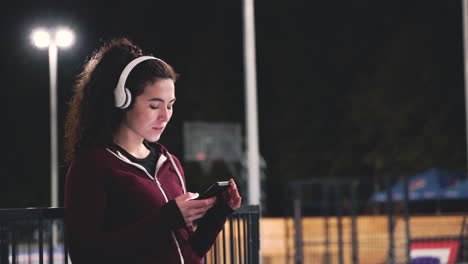 sportive curly woman listening music with bluetooth headphones and texting on her mobile phone while taking a break during her training session at night in the park