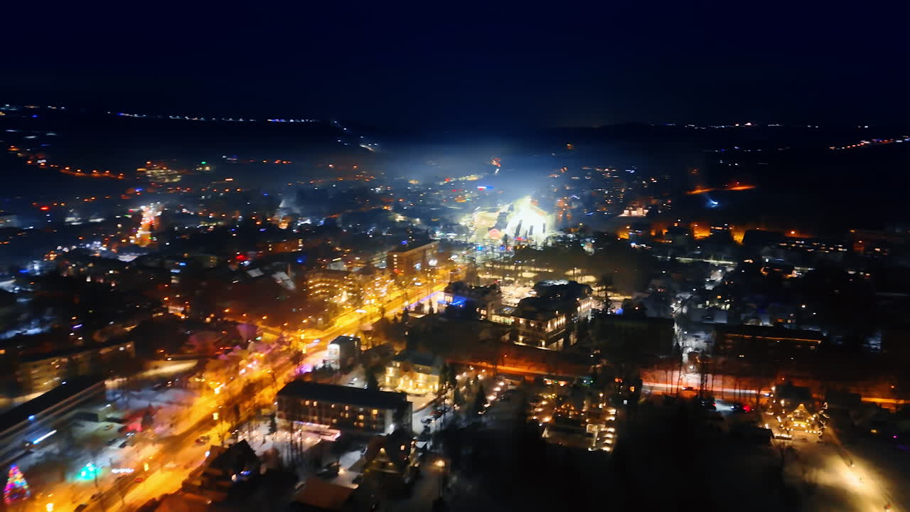 Avenue illuminated by the soft yellow light crosses the cozy town. Panorama of night Zakopane, Poland in winter. Aerial view.