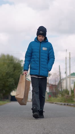 niño joven en chaqueta azul y gorra negra camina por una calle suburbana llevando una bolsa de papel, regresando de las compras, con casas residenciales y árboles de otoño alineando el camino