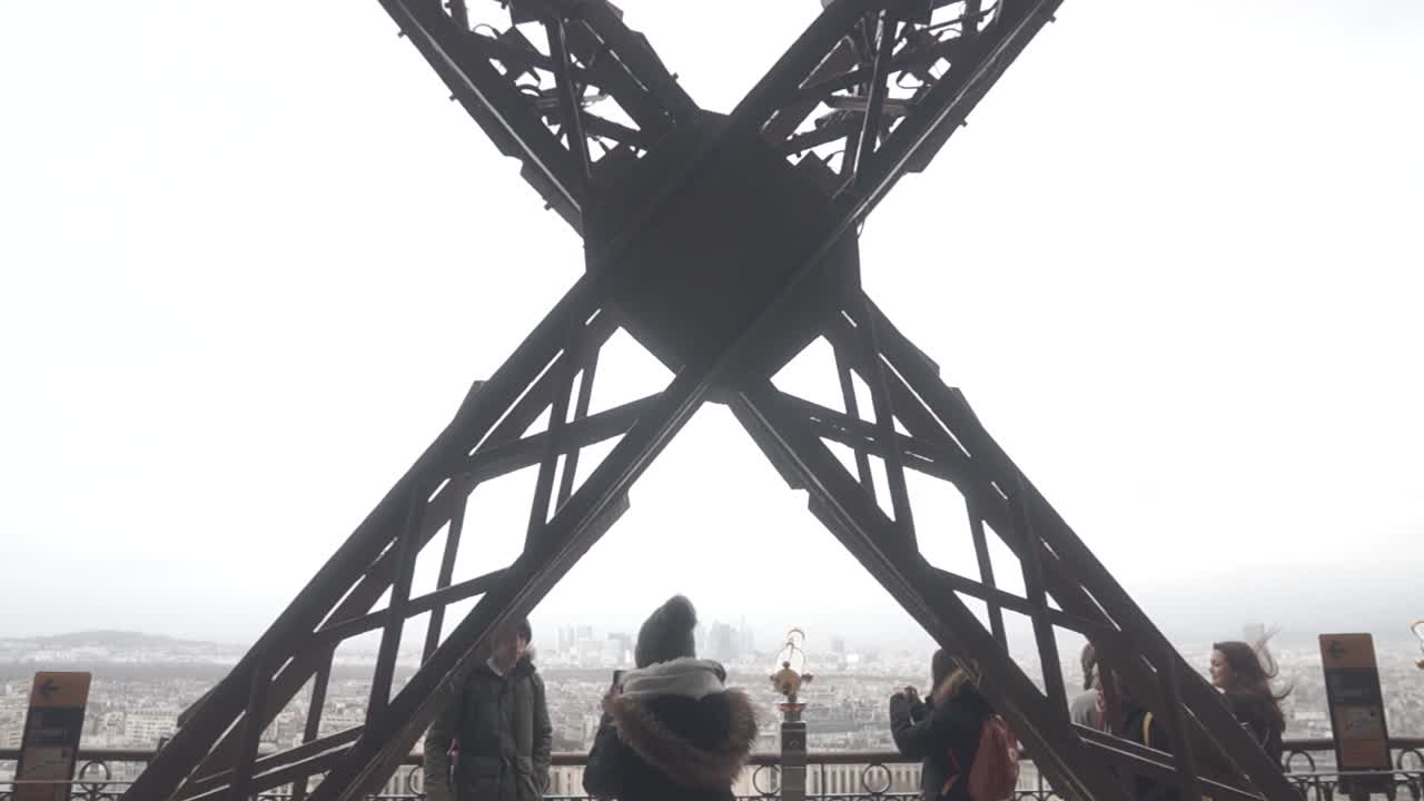 A push in on the trusses of the first level of the Eiffel Tower.  People are looking at the tower and the city.