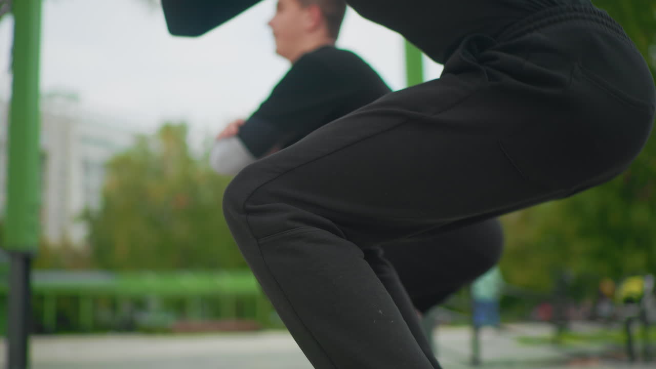 park squat intensity focused athlete, kontrollerede, lave squats med stram knæjustering, to træningspartnere over for metalrækværk, byparkbaggrund med bløde træer, matchende sorte leggings