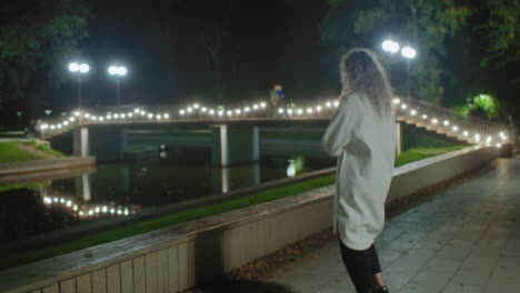 girl in white coat calmly approaches fence by water at night, crosses legs, opens book to read, while others stroll across lit bridge above tranquil reflection, fostering quiet study vibe in open air