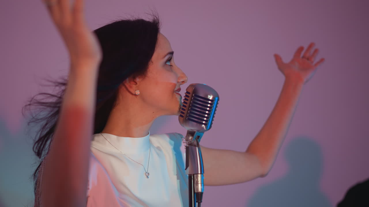 색 드레스를 입은 여성 가수가 빈티지 마이크 앞에 서서 손을 표현적으로 움직여 열정적으로 노래하고 있습니다. 기타리스트가 배경에서 연주합니다.