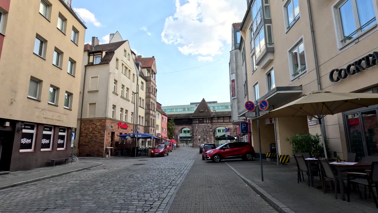 Camera glides along cobblestone street past colorful cafes toward historic city wall in daylight