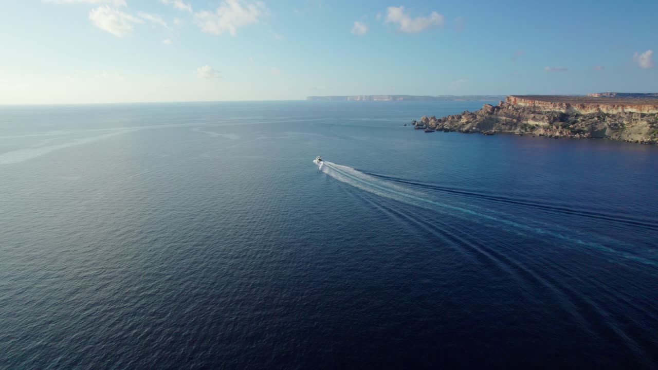 High aerial view over calm blue sea near Golden Bay, Malta, showing a distant boat leaving a long wake with rocky cliffs under a bright sky