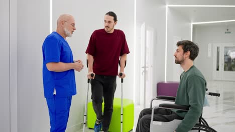 Un-Hombre-Seguro-De-Sí-Mismo-Con-Barba-Gris,-Un-Médico-Con-Uniforme-Azul-Se-Comunica-Con-Hombres-Después-De-Sus-Lesiones-En-Una-Clínica-Moderna-Con-Muletas-Y-Sillas-De-Ruedas-Para-Discapacitados