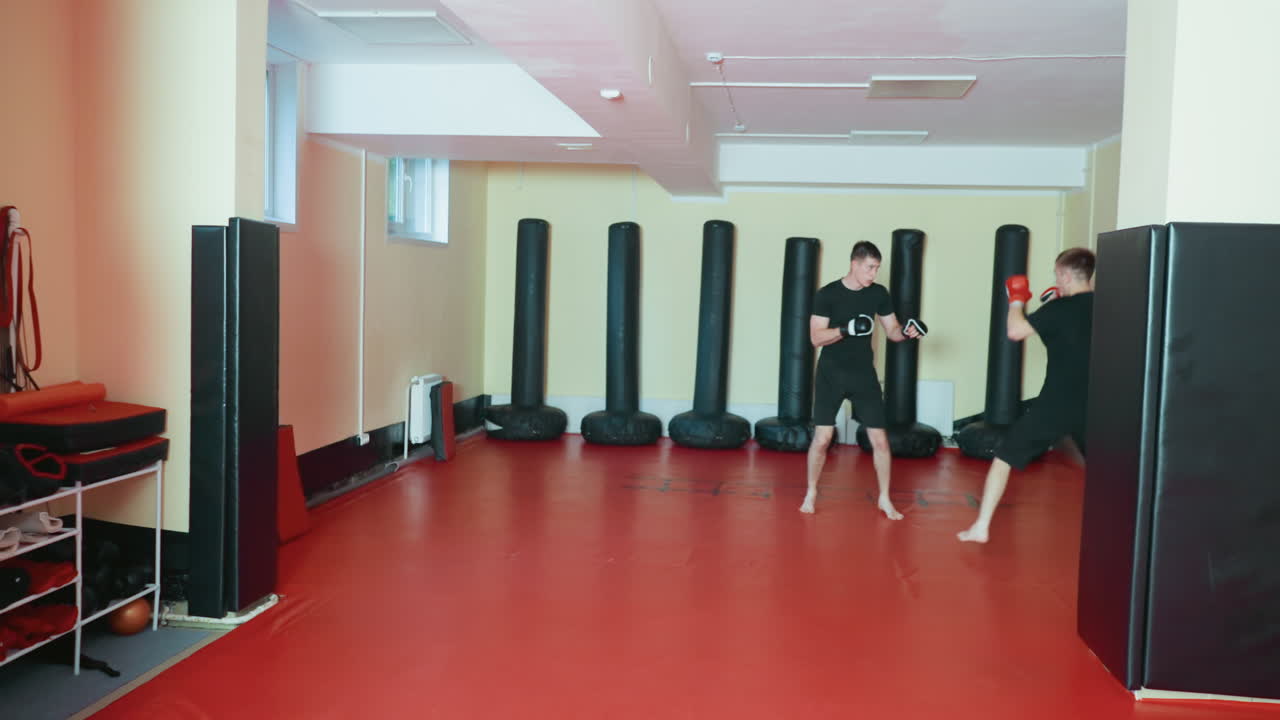 Two wrestlers spar barefoot on red mat in gym, wearing black sportswear and gloves, facing each other in defensive stances during combat practice, with punching bags lined against yellow wall