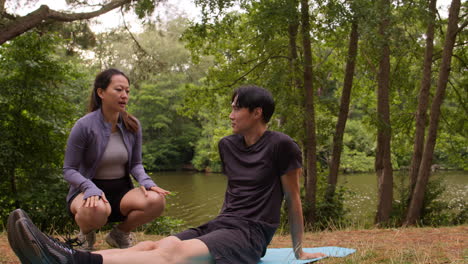 mujer enseñando a un hombre con ropa deportiva en una clase de yoga al aire libre rodeada de bosque y lago o río