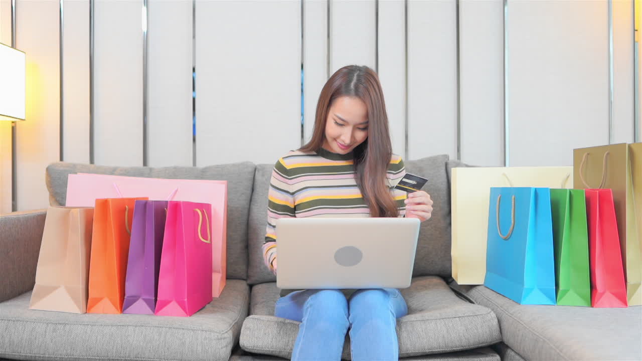 mujer asiática rodeada de coloridas bolsas de compras con computadora portátil y tarjeta de crédito comprando regalos en línea