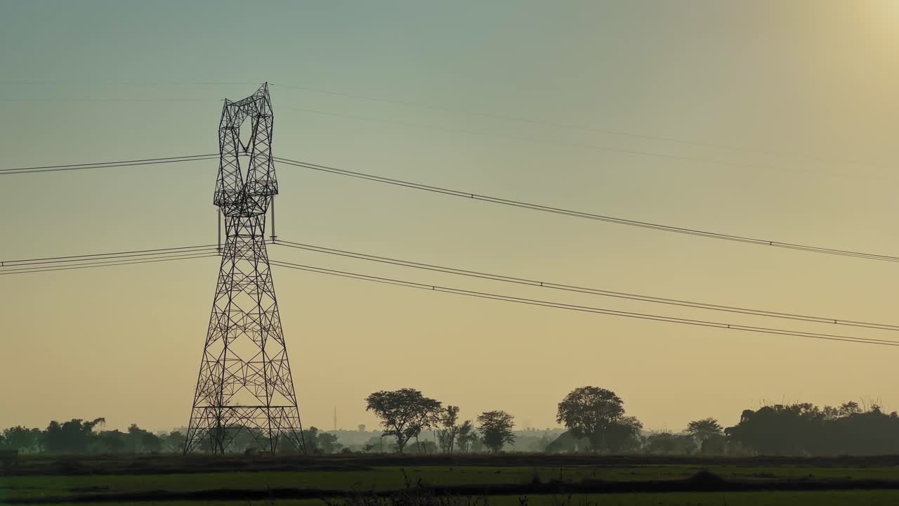 Steel lattice transmission tower carrying high voltage power lines across green agricultural fields under warm evening sky, showing contrast between energy infrastructure and rural landscape