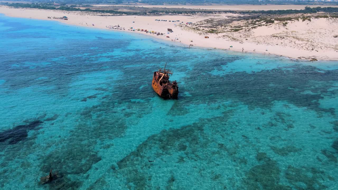 A rusted shipwreck semi-submerged in sparkling turquoise waters off the coastline of Tunisia, with a vibrant beach scene and desert horizon in the background.