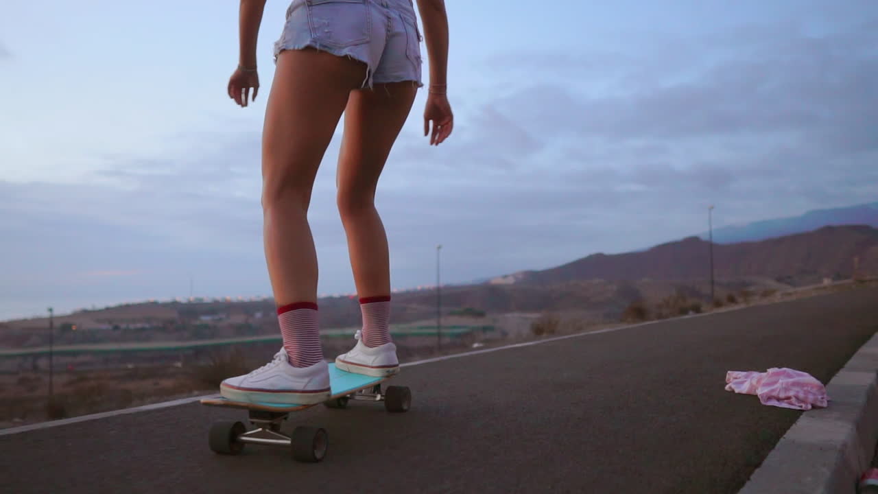 At sunset, a stunning and stylish skateboarder, sporting shorts, rides her board along a mountain road, and in slow motion, the mountains' view adds to the awe-inspiring scene