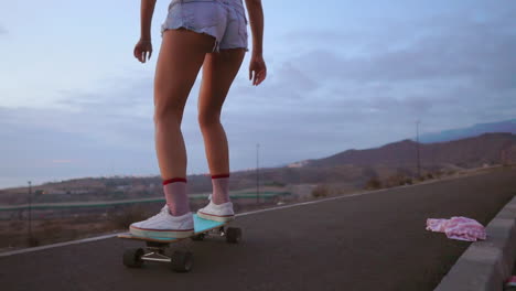 at sunset, a stunning and stylish skateboarder, sporting shorts, rides her board along a mountain road, and in slow motion, the mountains' view adds to the awe-inspiring scene