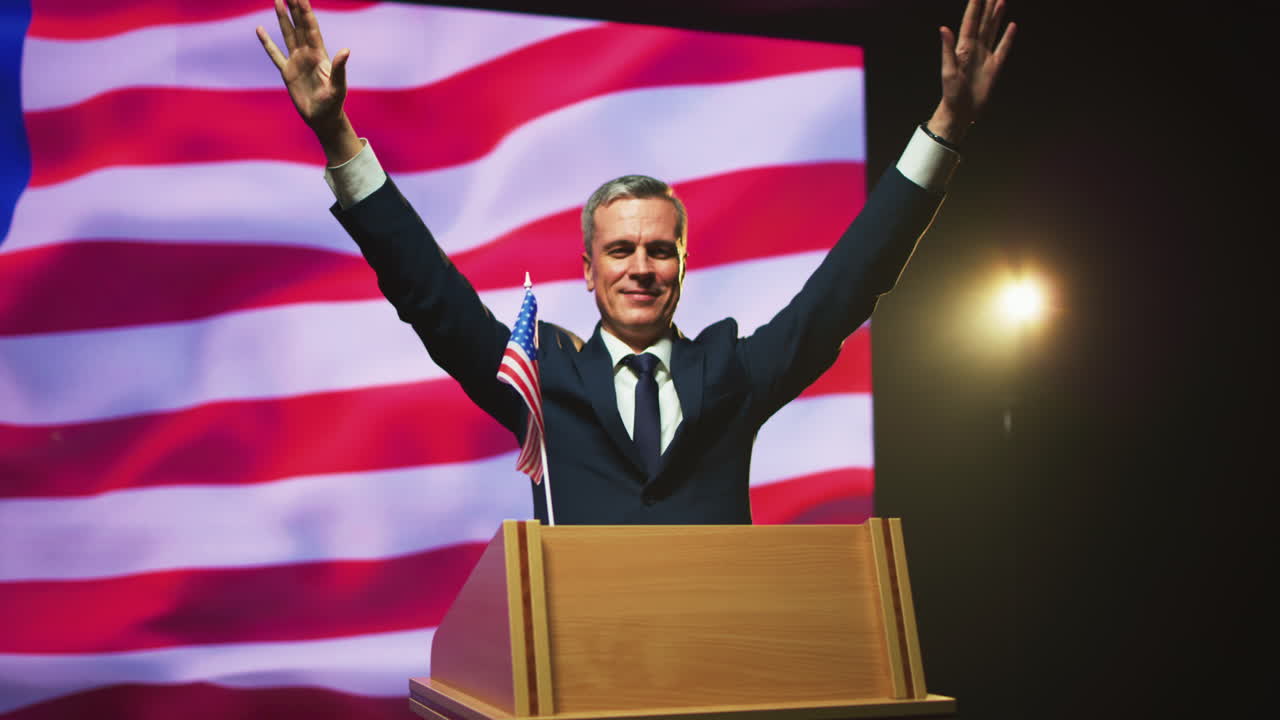 Happy Politician Gesticulating to Audience during Election Debate Zoom out View of Mature Man in Suit Gesticulating and Smiling to Audience while Standing on Rostrum under Photo Camera Flashes against Usa Flag after Election Speech