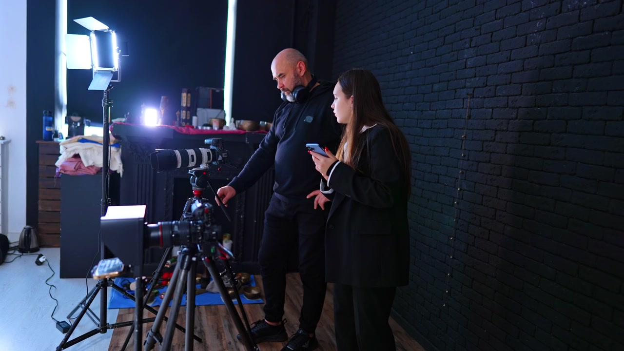 Videograf og assistent opsætter kamera i studie. Mand og assistent justerer kamera på stativ nær LED-lys og sort murstensvæg i studie