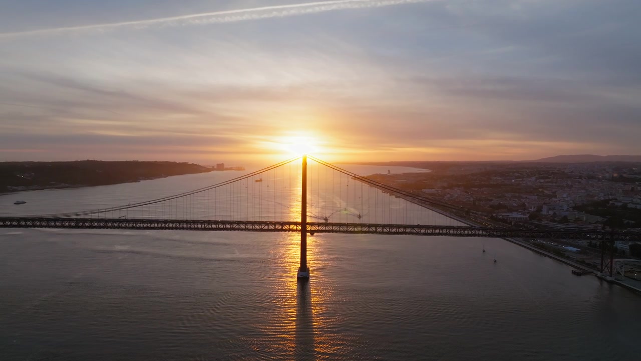 Riprese aeree catturano il Ponte 25 Aprile di Lisbona che attraversa il fiume Tago all'ora d'oro, con navi che scivolano sulle acque riflettenti e un tramonto vibrante che illumina lo skyline della città.