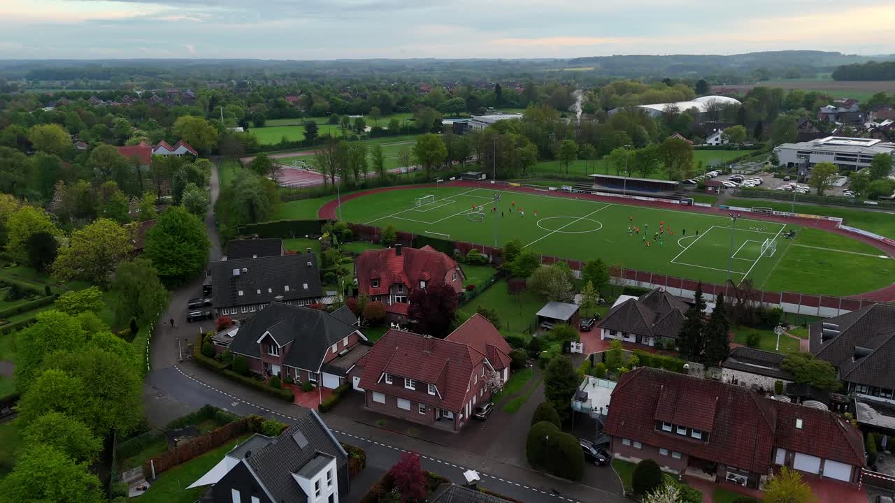 Running kids playing soccer on field in german neighborhood. Sunset time with clouds at sky in spring. Houses and homes in small city. Aerial forward wide shot.
