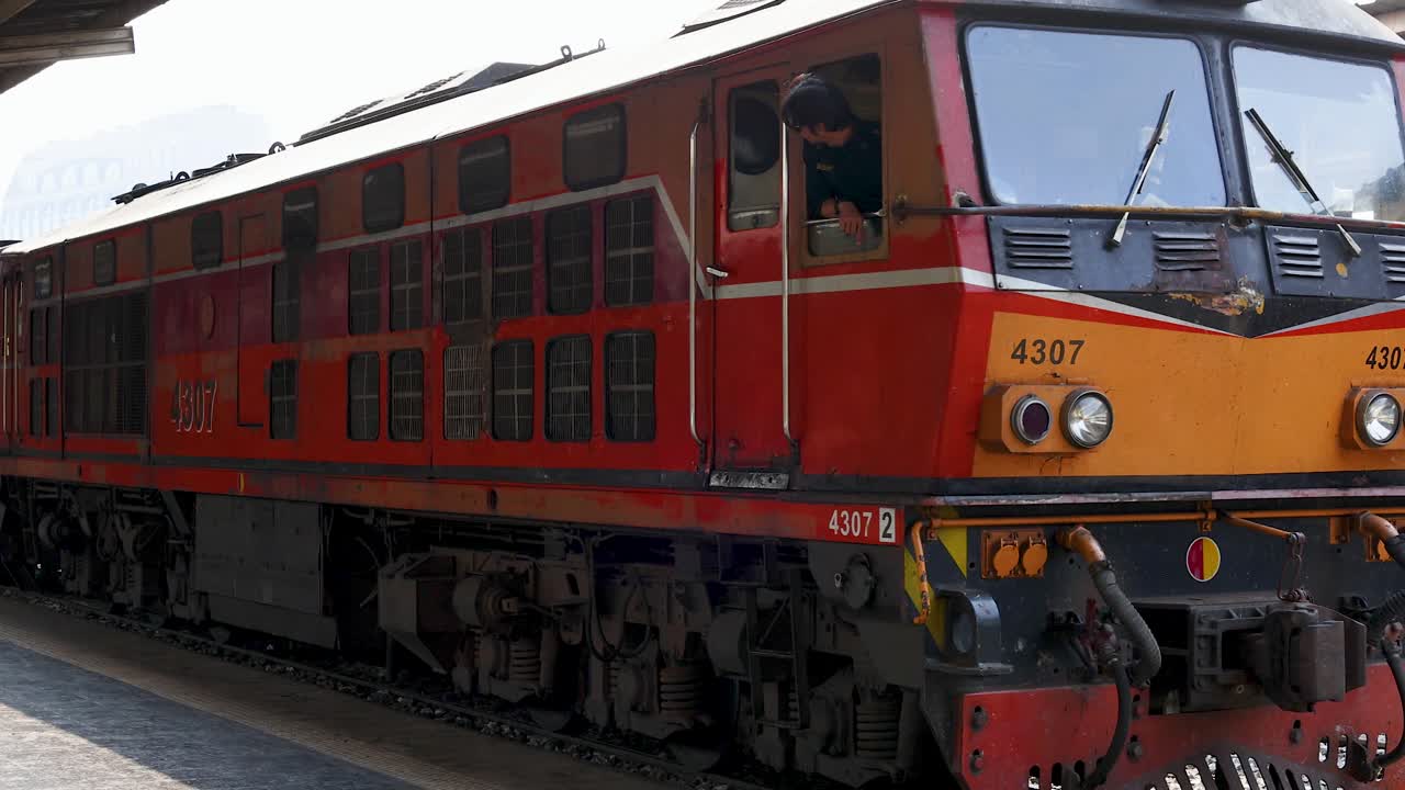 Slow-moving orange diesel locomotive passes platform at Bangkok station, daylight, handheld tracking shot