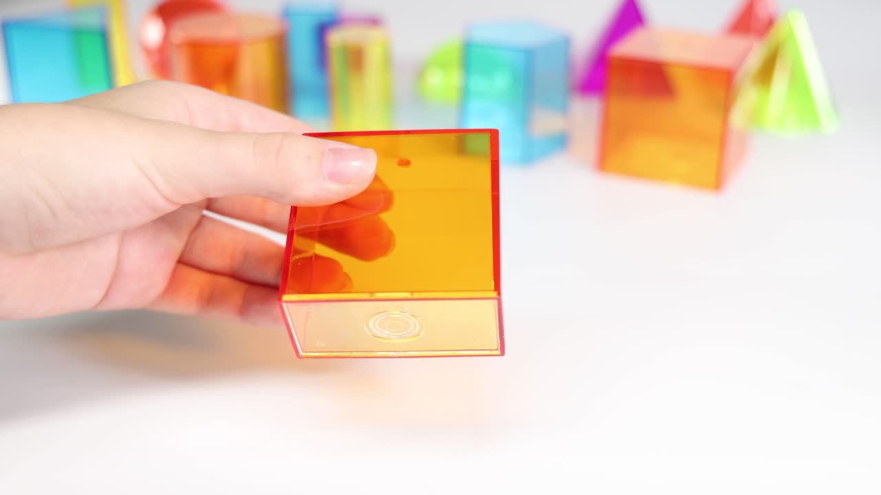 Hands manipulate translucent geometric shapes on a white surface. Bright colors and playful arrangement create an educational and engaging atmosphere