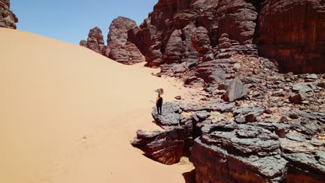 Tourist-Man-Standing-On-Edge-Of-A-Cliff-In-Djanet-Desert-In-Algeria---drone-shot