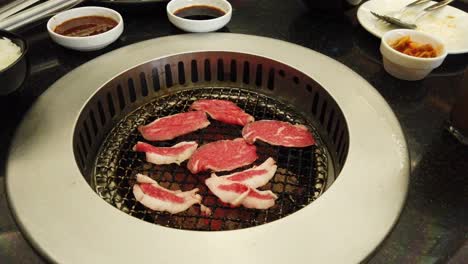 Fresh-and-thinly-sliced-beef-already-on-the-grill,-charcoal-burning,-condiments-in-small-saucers-around-an-actively-hot-grill