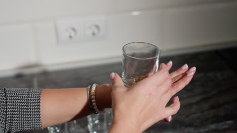 hand view of person with well polished nails picking glass cup from countertop placing it on back of hand playfully near scattered beads with clean marble surface and power outlet in kitchen setting