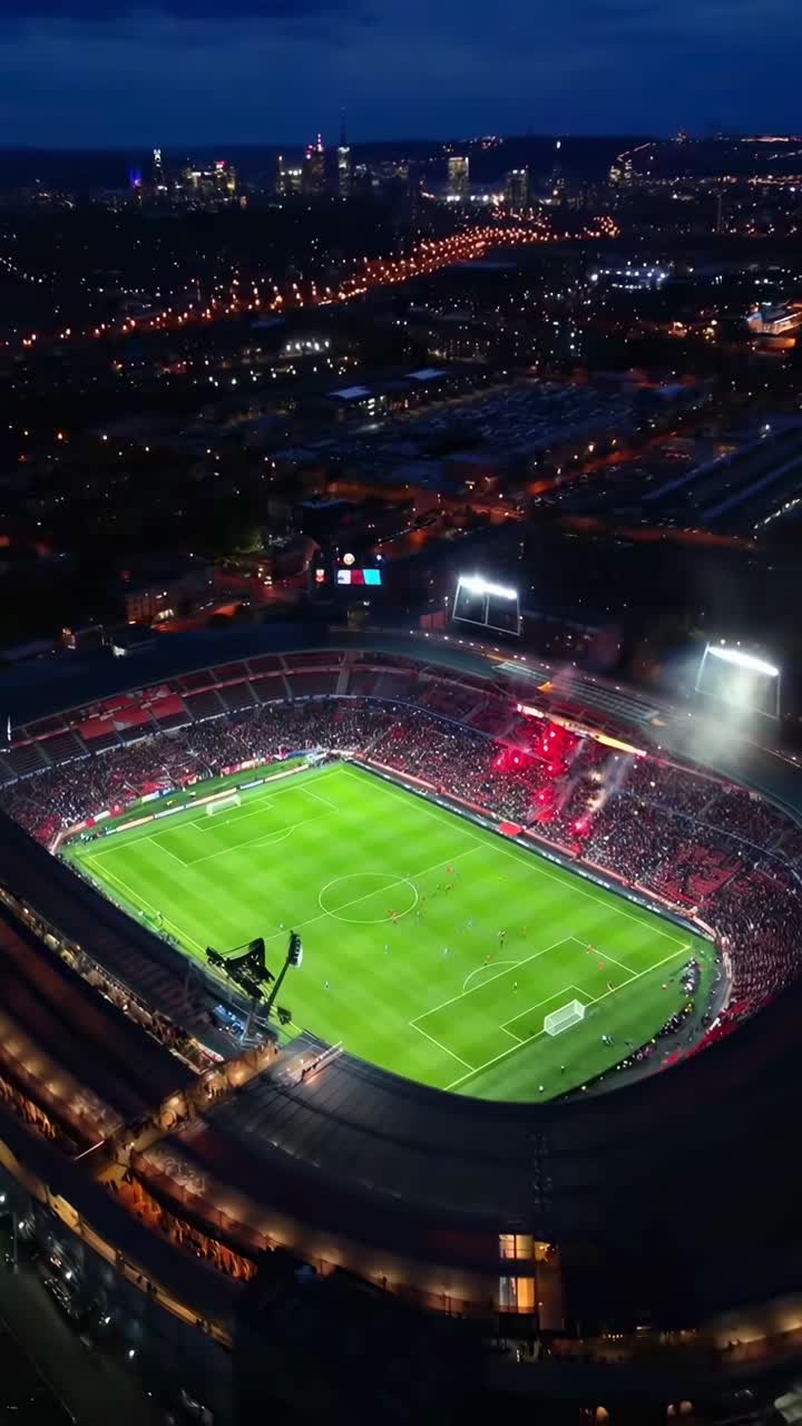 Un video aereo riprende uno stadio illuminato e vivace di notte, mostrando un evento sportivo animato