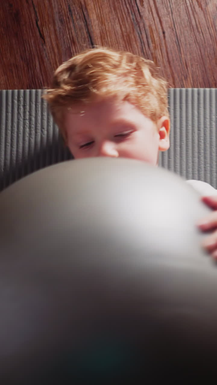 Little boy lies down on soft mat put on wooden floor holding silver gymnastic ball in hands. Cute toddler enjoys fitness training in home gym slow motion