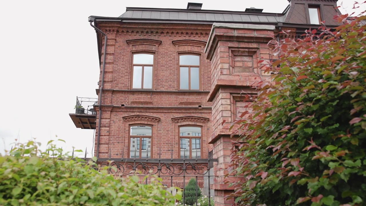 Un delicato movimento obliquo rivela una storica facciata residenziale in mattoni rossi con finestre simmetriche ad arco con cornice in legno, cornice a dentelli decorativa, stretto balcone metallico, piante in vaso e morbida vegetazione fogliosa.