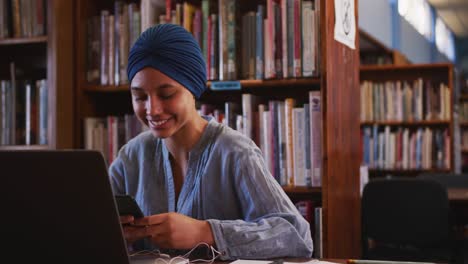 Asian-female-student-wearing-a-blue-hijab-sitting-with-a-laptop-and-using-a-smartphone