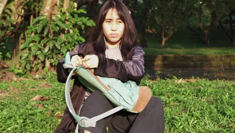 medium shot of asian female teenager sitting down at public park grass while opening backpack under bright sun