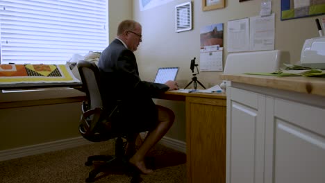 Mature-man-working-at-home-doing-video-conferencing-and-wearing-a-suit-jacket-and-tie-from-the-waist-up-but-shorts-and-bare-legs-those-on-the-conference-call-can't-see