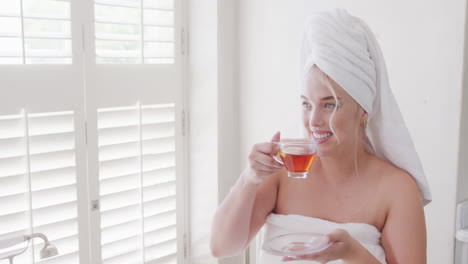 Happy-caucasian-plus-size-woman-with-towel-on-head-drinking-tea-in-slow-motion