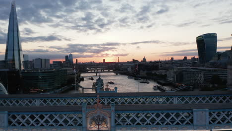 Backwards-fly-above-River-Thames-flowing-through-city-in-sunset-time.-Revealing-famous-Tower-Bridge.-London,-UK