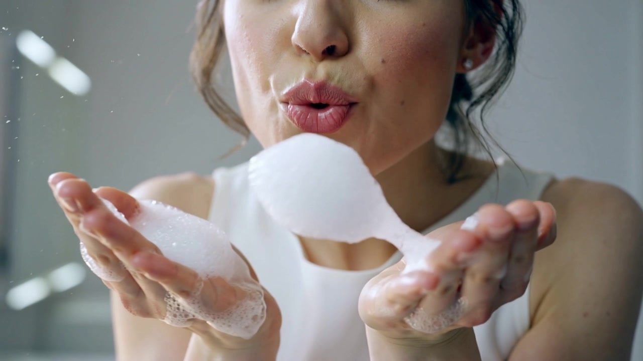 Donna che raccoglie la schiuma con le mani, stringe le labbra e soffia una bolla di schiuma davanti al lavabo del bagno, indossando un top senza maniche.