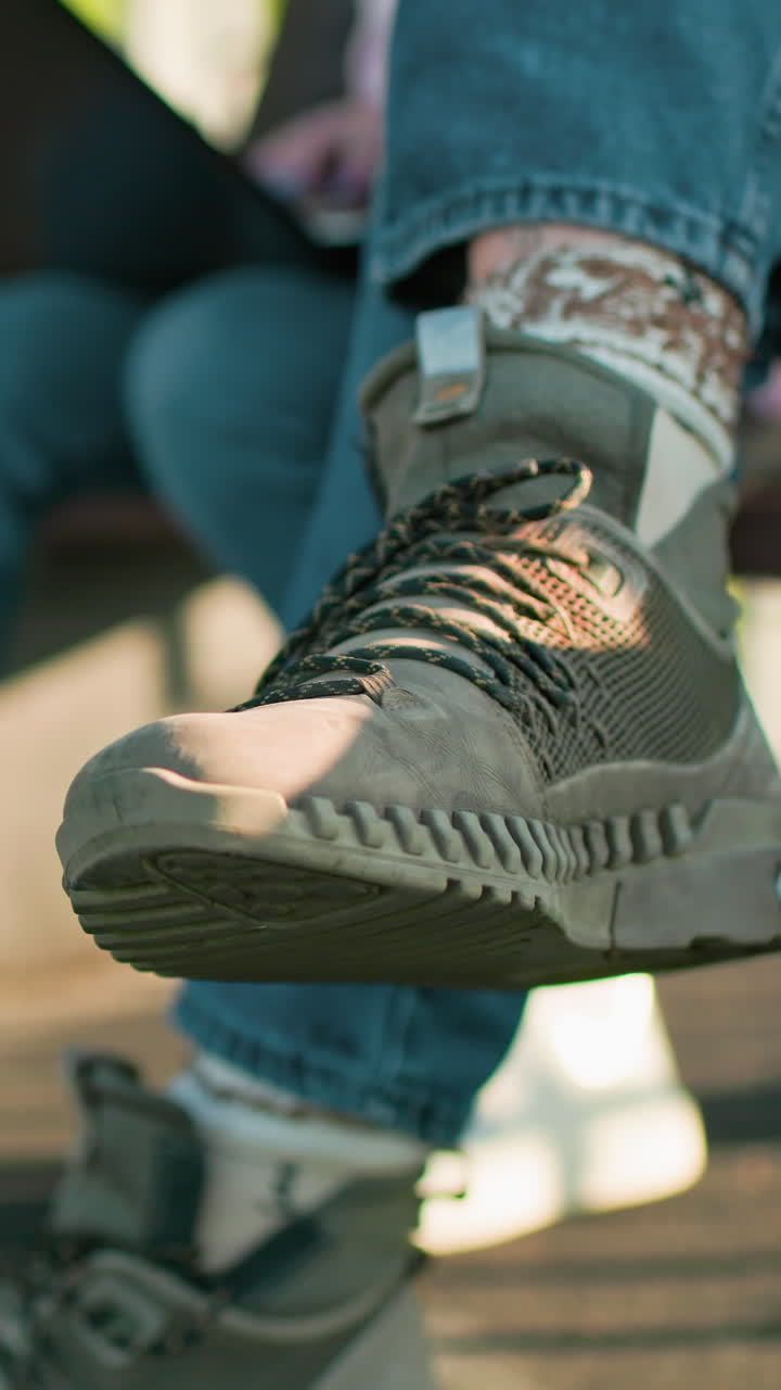 primer plano de las piernas de un hombre y una mujer sentados al aire libre en bancos de madera, con zapatillas deportivas y vaqueros casuales, uno usando una computadora portátil mientras la luz del sol proyecta sombras a través del entorno al aire libre relajado