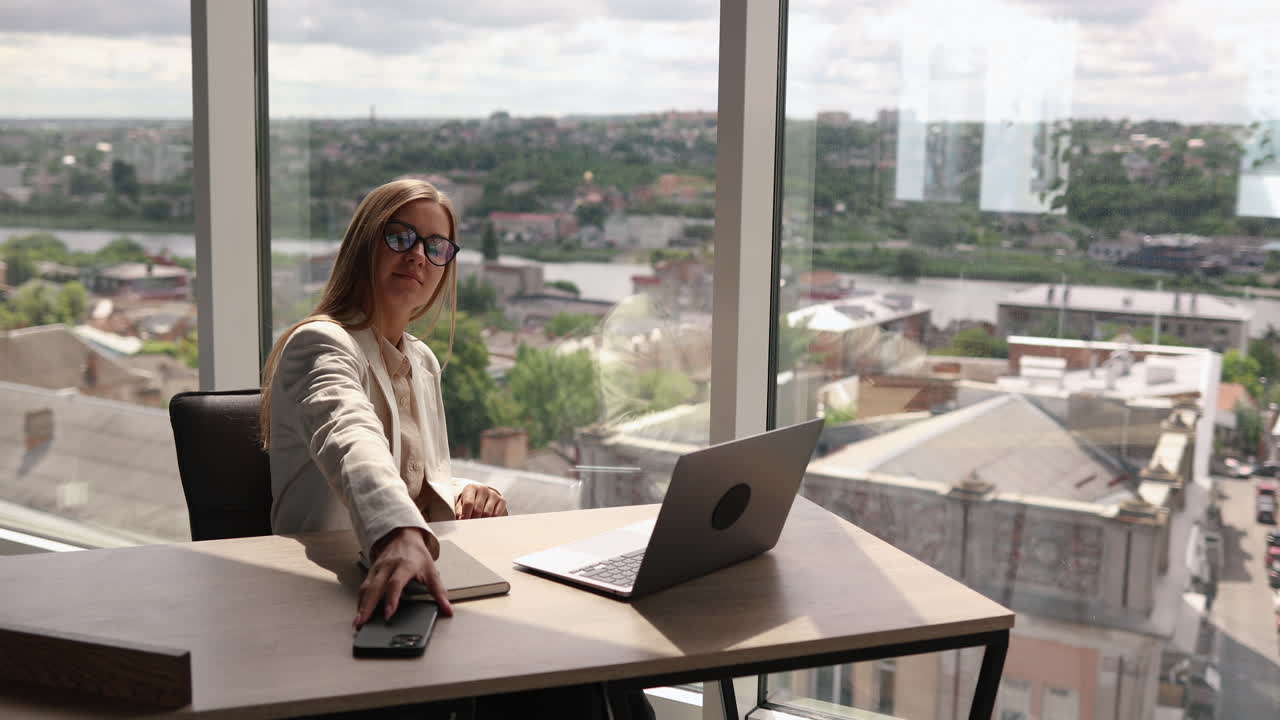 Una giovane donna di successo con gli occhiali siede rilassata alla sua scrivania, a fine giornata lavorativa in ufficio. Indossa abiti da lavoro classici.