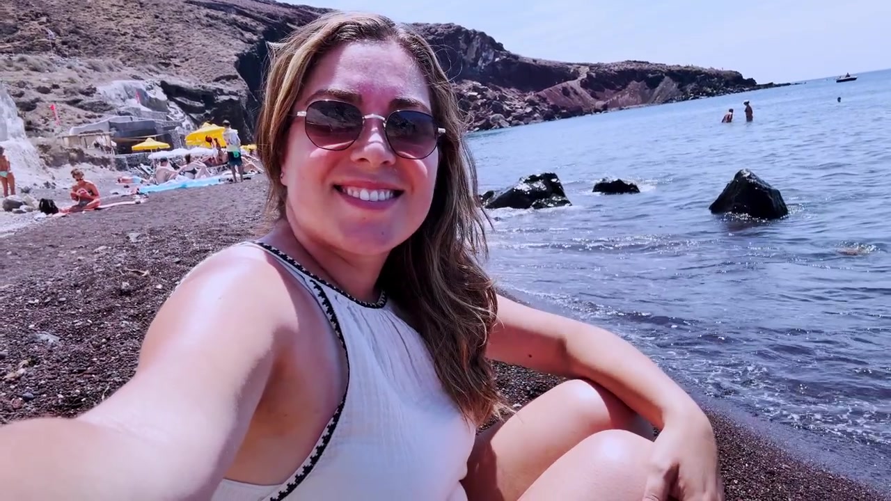 Iloinen vloggaaja kuvaamassa selfie-videota punaisella rannalla Santorinilla, Kreikassa.