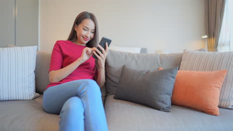 Asian-woman-playing-mobile-game-on-her-smartphone-sitting-on-comfortable-coach-with-many-pillow-at-home